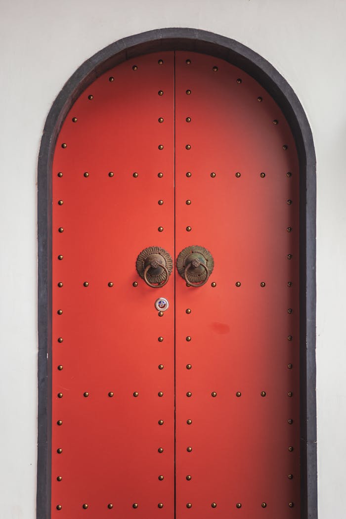 A striking red arched door with ornate metal knockers on a white background.