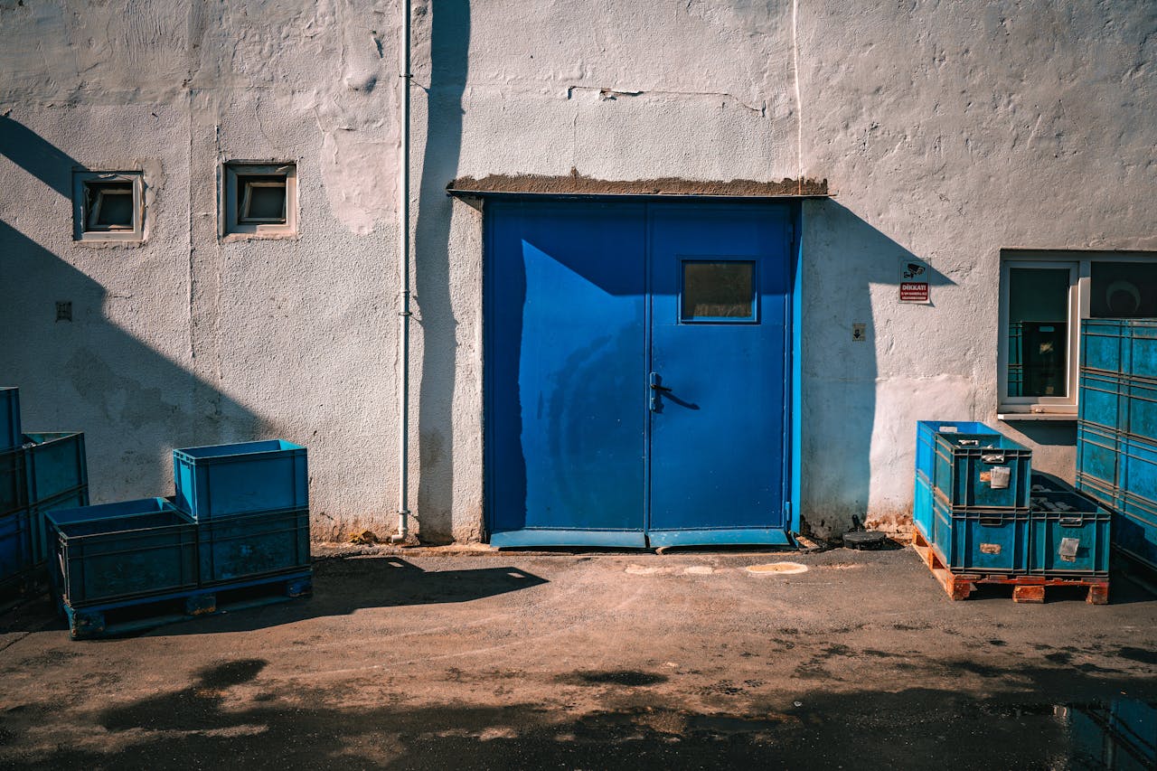 about-01 An industrial blue door surrounded by blue storage crates on a sunny day.